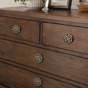 May include: A dark brown wooden dresser with decorative brass knobs. The knobs have an intricate, woven design. The dresser has multiple drawers and a smooth, polished surface. The overall style is classic and elegant.