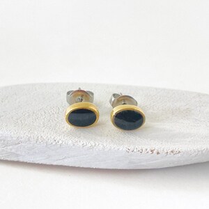 May include: A pair of oval stud earrings. Each earring features a black oval stone set in a gold-colored bezel. The posts are silver-colored. The earrings are displayed on a white, textured surface.
