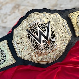 Puede incluir: Un cinturón de lucha de campeón mundial dorado y negro con el logotipo de la WWE. El cinturón tiene una placa central grande y dos placas laterales más pequeñas, todas con diseños intrincados y las palabras "World Champion".