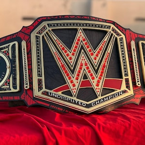 May include: A wrestling championship belt in black and red. The belt displays the WWE logo and the words "UNDISPUTED CHAMPION". Side plates are inscribed with "THE TRIBAL CHIEF". The belt is displayed on a red surface.