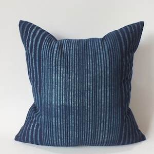 May include: A square decorative pillow with a cat-ear design. The pillow features a navy blue and light blue vertical striped pattern. The pillow is made of fabric and is placed against a white background.