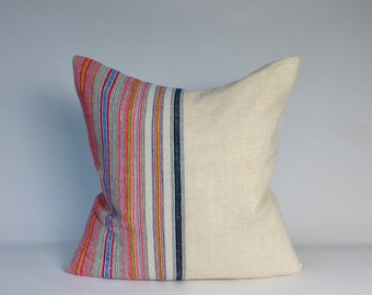 May include: A square decorative pillow with a two-tone design. The left side features vertical stripes in red, pink, blue, yellow, and white. The right side is a solid off-white color. The pillow is likely made of fabric.
