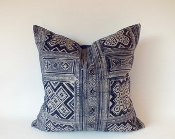 May include: Square pillow with a navy blue and off-white geometric pattern. The pillow features a repeating design of rectangles, squares, and other shapes. The pillow is likely made of fabric and is a decorative item for a bed or couch.