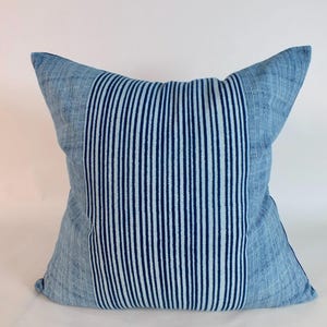 May include: A square throw cushion featuring a central vertical panel of dark blue and light blue stripes, flanked by side panels of a lighter, textured blue fabric with a subtle cross-hatched pattern throughout the entire surface of the cushion.