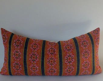 May include: Rectangular decorative pillow with a vibrant, patterned design. The pillow features a red background with vertical stripes of dark blue and intricate geometric patterns in yellow, pink, and white.