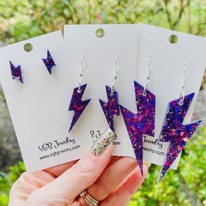 May include: Three pairs of purple and pink glitter lightning bolt earrings on white card stock with the text "VGB Jewelry www.vgbjewelry.com" on the left card and "Jewelry wetry.com" on the right card.