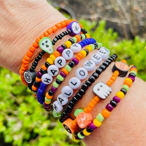 May include: A stack of seven beaded bracelets featuring orange, black, purple, green, and yellow seed beads. The bracelets include letter beads spelling out HAPPY HALLOWEEN, along with charms shaped like a skull, spider, ghost, pumpkin, and BOO text.