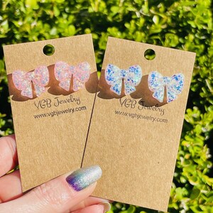 May include: Two pairs of bow-shaped stud earrings on brown cards. One pair is pink and white glitter, the other is blue and white glitter. The cards have the text "VGB Jewelry" and "www.vgbjewelry.com".