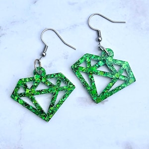 May include: A pair of green glitter diamond-shaped earrings with silver-colored ear wires. The earrings are made of a translucent green material with embedded glitter, creating a sparkling effect. The design is geometric and modern.