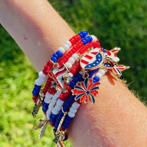 May include: A stack of beaded bracelets in red, white, and blue, with gold-colored charms. The charms include stars, fireworks, and a shield design. The bracelets are worn on a person's arm.