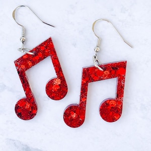 May include: A pair of red glitter musical note earrings. The earrings are shaped like eighth notes with a silver hook. The red glitter is visible throughout the earrings. The earrings are on a white background.