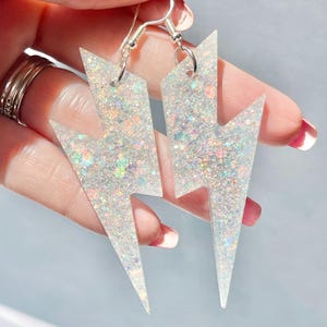 May include: A pair of lightning bolt shaped dangle earrings featuring a clear resin base filled with iridescent, multi-coloured glitter. The earrings are attached to silver-toned metal fishhook style ear wires for hanging.
