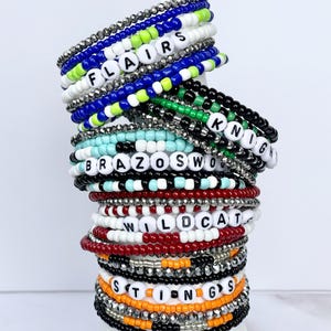 May include: A stack of beaded bracelets in various colors, including white, blue, black, and red. Some bracelets feature white letter beads spelling out words like "FLAIRS," "KNIGHT," and "WILDCAT." The bracelets are stacked on top of each other.