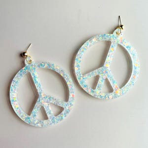 May include: A pair of glittery peace sign earrings. The earrings are a light blue color with iridescent glitter throughout. The earrings have gold-colored ear wires.