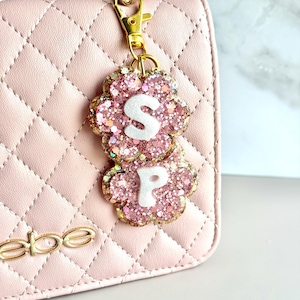 Peut inclure: Un porte-clés en métal doré composé de deux breloques en forme de fleur remplies de paillettes roses. La fleur supérieure affiche une lettre S blanche et la fleur inférieure une lettre P blanche. Le porte-clés est fixé à un sac matelassé rose pâle.