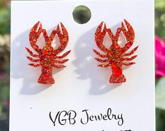 May include: A pair of red glitter lobster stud earrings. The earrings are on a white card with the text "VGB Jewelry" and the website address. The earrings are a bright, eye-catching accessory.