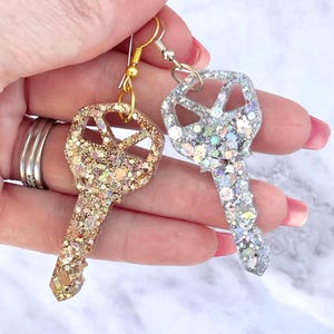 May include: Two key-shaped earrings. One is gold-colored with gold glitter, the other is silver-colored with silver glitter. Both earrings have a fish hook style ear wire.