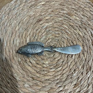 May include: A silver-colored fish-shaped butter knife with the word "mudpie" engraved on the blade. The knife is lying on a woven, circular placemat made of natural fibers. The fish has detailed scales and fins.
