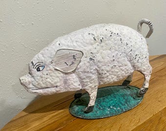 May include: A vintage metal pig figurine with a textured white body and a curled tail. The pig stands on a green oval base. The figurine has black hooves and is placed on a wooden surface.