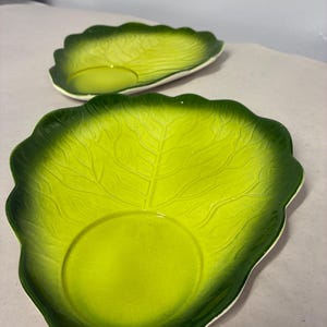 May include: Two ceramic dishes shaped like cabbage leaves, featuring a vibrant gradient of lime green in the center transitioning to a deep forest green along the scalloped, textured edges. Each dish has a smooth, circular indentation in the middle.