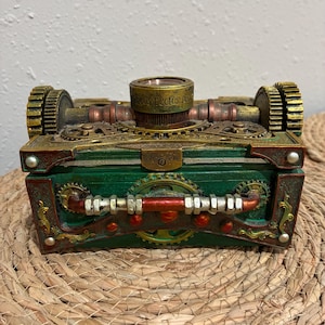 May include: A steampunk-style box with a green base, adorned with bronze gears, copper pipes, and gold accents. The box features a handle and a circular top with the text "meus pectus prae".