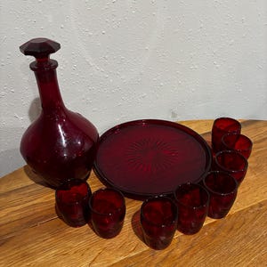 May include: A vintage ruby red glass decanter with a faceted stopper, a round tray, and eight matching shot glasses. The glassware is arranged on a wooden surface, showcasing the rich color and classic design.