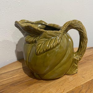 May include: Green ceramic pitcher with a leaf-shaped rim and handle. The pitcher has a textured surface and a bulbous body. The handle is designed to look like a tree branch. The pitcher is sitting on a wooden surface.