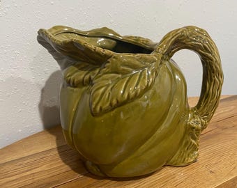 May include: Green ceramic pitcher with a leaf-shaped rim and handle. The pitcher has a textured surface and a bulbous body. The handle is designed to look like a tree branch. The pitcher is sitting on a wooden surface.