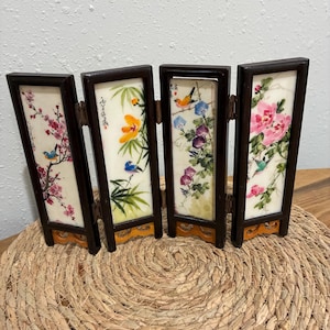 May include: A four-panel folding screen with floral and bird illustrations. Each panel features a different design: cherry blossoms, bamboo with an orange flower, morning glories, and pink peonies. The frames are dark brown, and the bases are a light brown color.