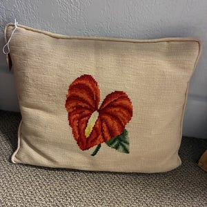 May include: A rectangular needlepoint pillow featuring a beige canvas background with a centered, vibrant red anthurium flower design. The flower has a prominent yellow spadix and a single green leaf at the base, finished with matching beige piping.