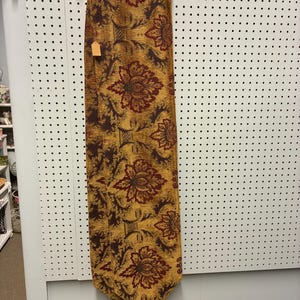 May include: A rectangular table runner featuring a woven mustard yellow background with a repeating pattern of dark red floral motifs and brown foliage. The runner is finished with two dark blue tassels attached to the bottom edge.