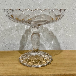 May include: A clear glass pedestal candy dish with a scalloped rim. The dish has a round base and a decorative stem. The dish is sitting on a wooden surface.