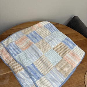 May include: A square patchwork quilt featuring a grid of fabric squares with various striped patterns in shades of light blue, tan, and soft peach. The quilt is finished with a solid light blue fabric border along the outer edges.