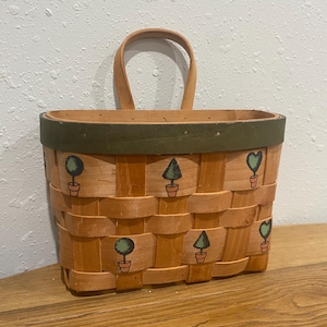 May include: Woven rectangular basket with a handle, featuring a light brown color with a green trim. The basket is decorated with painted green potted trees and hearts. The basket is sitting on a wooden surface.