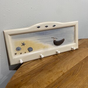 May include: A rectangular wall-mounted coat rack with a beach scene painted on it. The artwork features a boat, seashells, and a starfish. The rack has a cream-colored frame and six white knobs for hanging items.