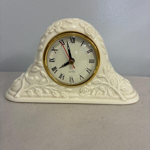 May include: An off-white mantel clock with a molded floral and leaf relief pattern. The clock face features a gold-tone circular frame, black Roman numerals, and black hour and minute hands with a red second hand. The text QUARTZ and TAIWAN are printed on the face.