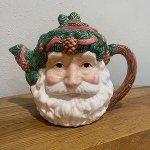 May include: A ceramic teapot shaped like Santa Claus's face. The teapot features a white beard, rosy cheeks, and a green and pink wreath on its head. The handle is brown, and the spout is on the left side. A festive holiday decoration.
