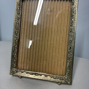 May include: A rectangular picture frame with an ornate, embossed gold-tone metal border featuring intricate scrollwork and decorative corner accents. The frame is supported by two small, rounded metal feet at the base and contains a clear glass pane.
