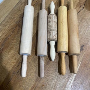 May include: Five wooden rolling pins of varying sizes and designs. The rolling pins are made of light-colored wood, with different handle shapes. One pin has a carved floral pattern. The rolling pins are arranged on a wooden surface.