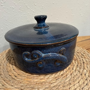 May include: A blue ceramic casserole dish with a lid. The dish has decorative handles and a dark blue glaze with brown accents. The dish is sitting on a woven placemat.