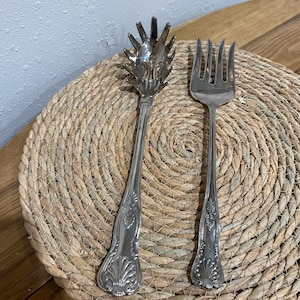 May include: Two silver serving utensils, a spaghetti server and a fork, rest on a woven placemat. The spaghetti server has a starburst head, while the fork has four tines. Both have ornate handle detailing.