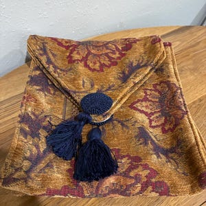 May include: A decorative clutch with a floral pattern in shades of gold, burgundy, and purple. The clutch features a flap closure and is adorned with a navy blue button and two navy blue tassels. The bag is made of a textured fabric.