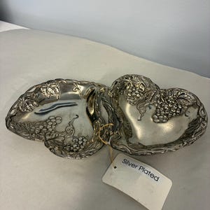 May include: A silver-plated double dish featuring two heart-shaped bowls connected together. The edges and surfaces are decorated with embossed grape clusters and vine leaf patterns. A white rectangular tag attached by a string reads Silver Plated.