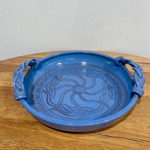 May include: A round, light blue ceramic serving bowl with two braided handles. The bowl features a detailed, sunburst-like pattern in the center. The bowl is displayed on a wooden surface.