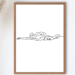 May include: A framed art print featuring a minimalist line drawing of a swimmer in the freestyle stroke. The artwork is in black against a white background, encased in a wooden frame. The print is labeled "aqualia print".