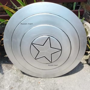 May include: A silver-colored shield with a central star design. The shield has concentric circular patterns and the text "Rozain mart" is visible on the surface. The shield is likely a prop or collectible.