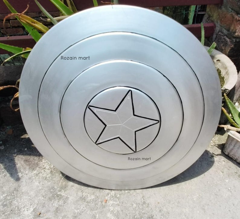 May include: A silver-colored shield with a central star design. The shield has concentric circular patterns and the text "Rozain mart" is visible on the surface. The shield is likely a prop or collectible.