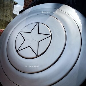 May include: A silver-colored shield with a central white star design, reminiscent of a superhero's emblem. The shield has concentric circular ridges and the text "Rozain mart" is visible on the surface. This item is likely a collectible or cosplay accessory.