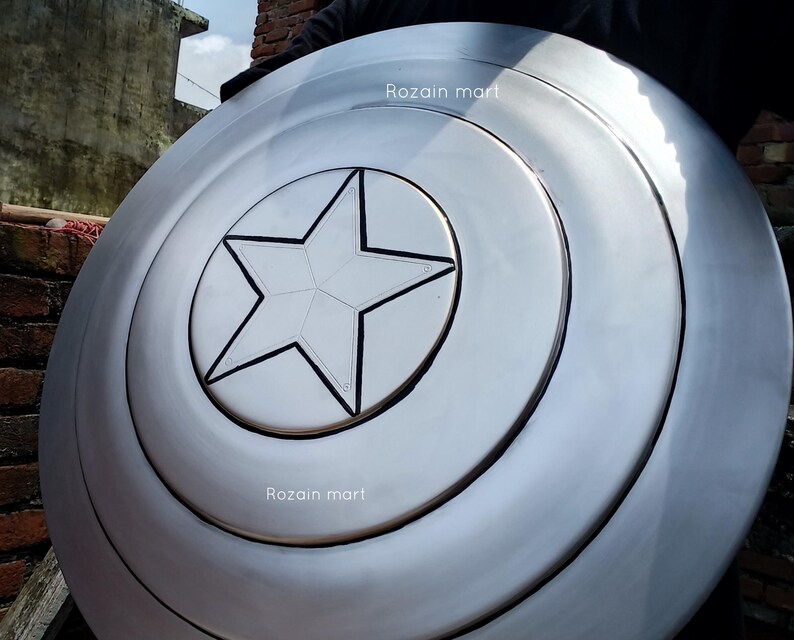 May include: A silver-colored shield with a central white star design, reminiscent of a superhero's emblem. The shield has concentric circular ridges and the text "Rozain mart" is visible on the surface. This item is likely a collectible or cosplay accessory.
