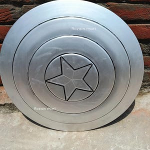 May include: A silver-colored, circular shield with a five-pointed star in the center. The shield has concentric rings and the text "Rozain mart" is visible on the surface. The shield is likely a prop or collectible.